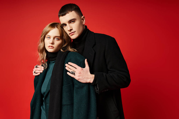 alluring loving couple in sophisticated chic coats looking at camera on red vibrant background - Fotó, kép