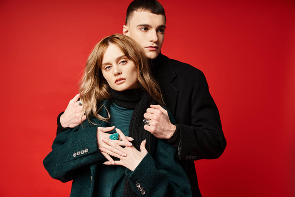 appealing woman in coat looking at camera and posing lovingly with her boyfriend on red backdrop - Foto, afbeelding