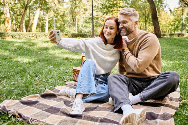 Άνδρας και γυναίκα στο πάρκο, κάθεται σε κουβέρτα, συλλαμβάνοντας στιγμή με selfie. - Φωτογραφία, εικόνα