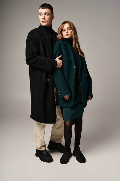 good looking stylish boyfriend and girlfriend in coats posing lovingly together on gray backdrop - Photo, Image