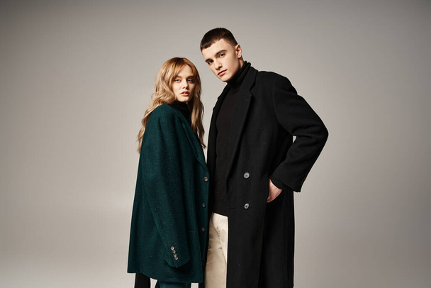 fashionable couple in stylish coats posing together on gray backdrop and looking at camera - Foto, Imagem