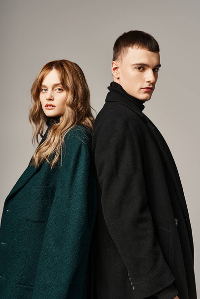 appealing trendy boyfriend and girlfriend in coats posing and looking at camera on gray backdrop - Фото, изображение