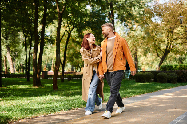 A man and woman in casual attire walk down a peaceful path in a lush park. - Photo, Image