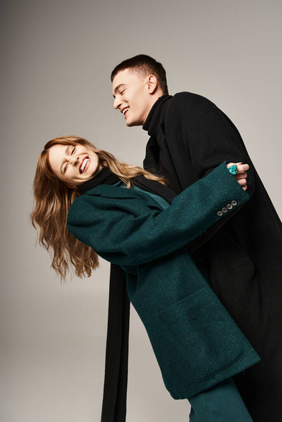 alegre casal atraente em casacos elegantes sorrindo alegremente e tendo tempo de qualidade em pano de fundo cinza - Foto, Imagem
