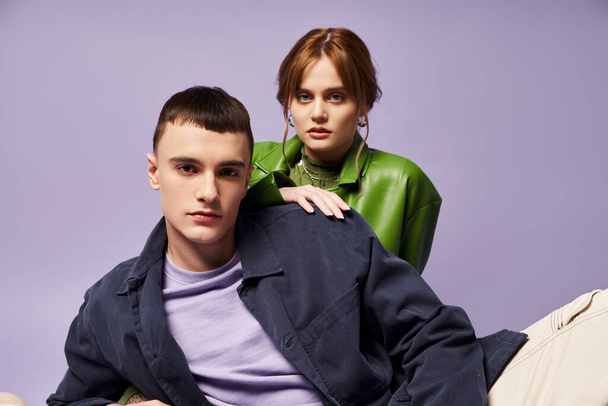 sophisticated couple in vibrant attires sitting on floor and looking at camera on purple backdrop - Фото, изображение