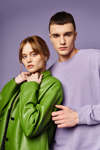 sofistikovaný pár v pulzujícím oblečení pózovat společně na fialovém pozadí a při pohledu na kameru - Fotografie, Obrázek