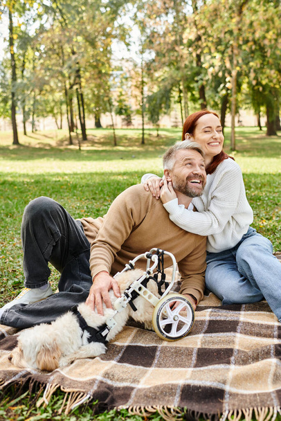 A loving couple relaxes on a blanket with their furry companion in a peaceful park setting. - Photo, Image
