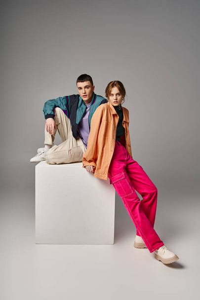 loving appealing couple in vivid stylish bombers looking at camera on gray backdrop on white cube - Photo, Image