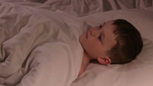 Chlapec s otevřenýma očima nemůže v noci spát v posteli v ložnici a snít. Dítě syn se dívá na strop na polštáři pod přikrývkou ve večerních hodinách - Záběry, video