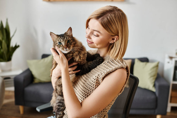 Eine Frau mit kurzen Haaren sitzt auf einem Stuhl und hält zu Hause friedlich eine flauschige Katze im Arm. - Foto, Bild