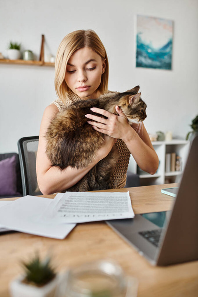 Μια γυναίκα με κοντά μαλλιά κάθεται σε ένα γραφείο, κρατώντας μια γάτα στην αγκαλιά της, ενώ στο σπίτι. - Φωτογραφία, εικόνα