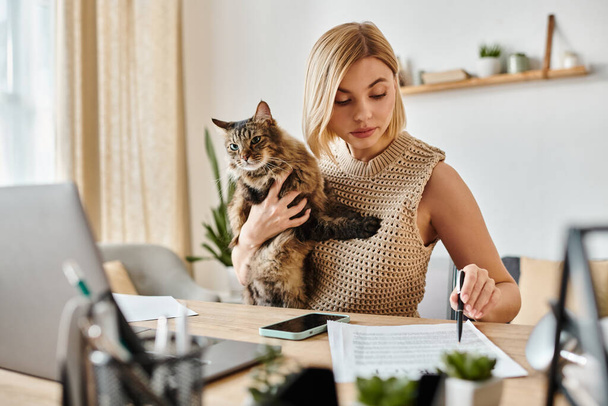 Μια γυναίκα με κοντά μαλλιά κάθεται σε ένα γραφείο στο εσωτερικό, κρατώντας μια γάτα περιεχόμενο στην αγκαλιά της, απολαμβάνοντας μια ειρηνική στιγμή στο σπίτι. - Φωτογραφία, εικόνα