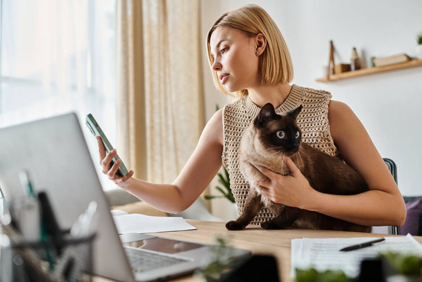 Μια γυναίκα με κοντά μαλλιά κρατά μια γάτα, ενώ επικεντρώνεται σε μια οθόνη laptop στο σπίτι. - Φωτογραφία, εικόνα