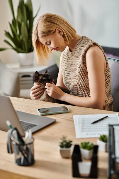Kobieta z krótkimi włosami relaksująca się przy biurku z kotem spokojnie zagnieżdżonym na kolanach. - Zdjęcie, obraz
