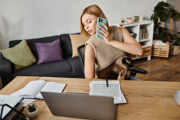 Egy rövid hajú, elegáns nő ül az asztalnál, a mobiljába merülve, miközben a macskája ott lóg mellette.. - Fotó, kép