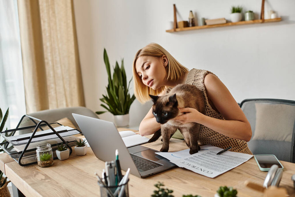 Μια ελκυστική γυναίκα με κοντά μαλλιά κάθεται σε ένα γραφείο, χαιδεύοντας με ικανοποίηση μια γάτα στην αγκαλιά της. - Φωτογραφία, εικόνα