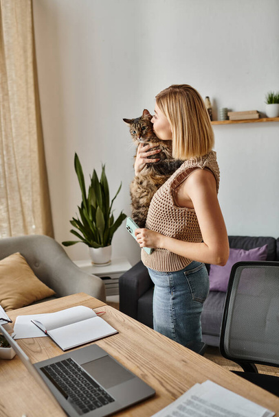 Spokojna kobieta z krótkimi włosami trzyma kota, stojącego przed laptopem w przytulnym domu. - Zdjęcie, obraz