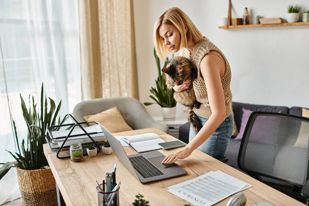A stylish woman stands at a desk, working on a laptop, as her loyal cat keeps her company with a playful presence. - Φωτογραφία, εικόνα