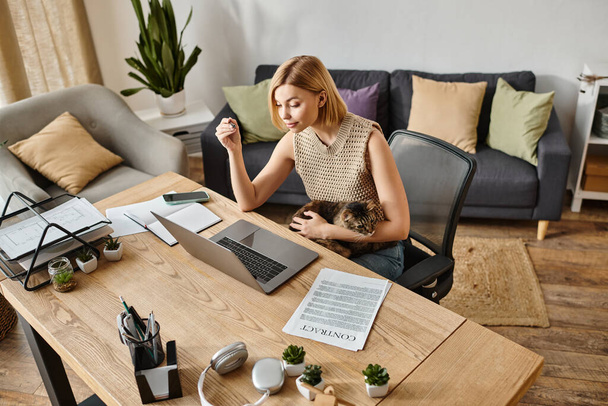 Uma mulher elegante com cabelo curto sentado em uma mesa com um laptop, acompanhado por seu gato em um ambiente acolhedor casa. - Foto, Imagem