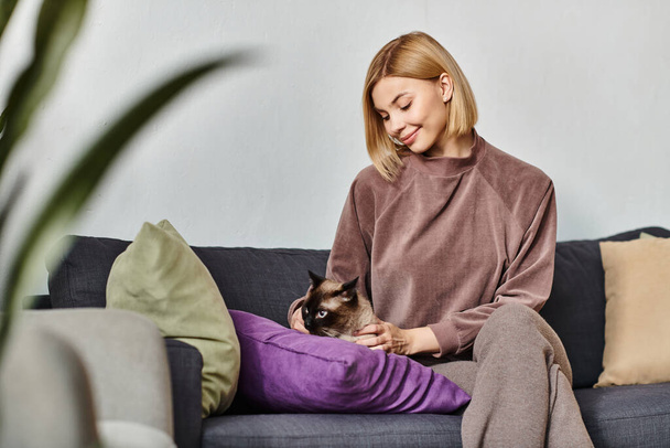 Μια γαλήνια γυναίκα με κοντά μαλλιά χαλαρώνει σε έναν καναπέ, κρατώντας απαλά την αγαπημένη της γάτα κοντά στο στήθος της. - Φωτογραφία, εικόνα