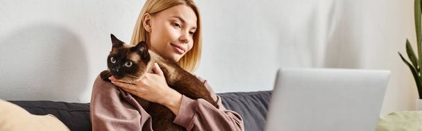 Μια γυναίκα με κοντά μαλλιά χαλαρώνει σε έναν καναπέ, κρατώντας ειρηνικά τη γάτα της σε μια ζεστή στιγμή στο σπίτι. - Φωτογραφία, εικόνα