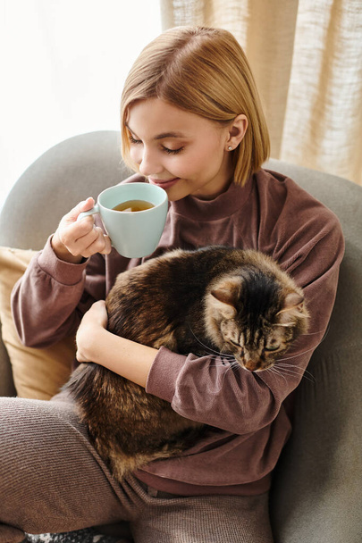 Egy rövid hajú nő, aki békésen ül egy székben, egy macskát tart az ölében, miközben egy csésze kávét iszik.. - Fotó, kép