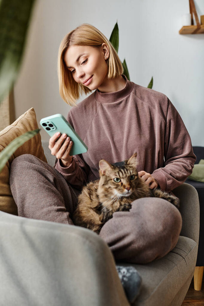 Μια ελκυστική γυναίκα με κοντά μαλλιά κάθεται σε έναν καναπέ, κρατώντας τη γάτα της σε μια ειρηνική και οικεία στιγμή στο σπίτι. - Φωτογραφία, εικόνα