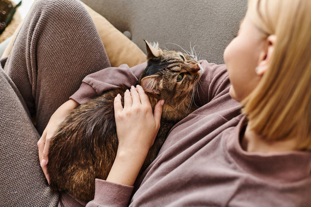 Kobieta z krótkimi włosami leżąca na kanapie, delikatnie pieszcząca kota spokojnym wyrazem twarzy. - Zdjęcie, obraz