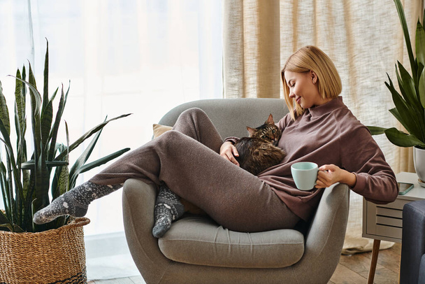 Kobieta z krótkimi włosami relaksuje się na krześle, gdy jej kot siedzi na kolanach.. - Zdjęcie, obraz