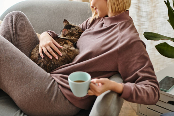 Μια γυναίκα με κοντά μαλλιά χαλαρώνει σε έναν καναπέ, κρατώντας ένα φλιτζάνι καφέ και μια γάτα περιεχόμενο στην αγκαλιά της. - Φωτογραφία, εικόνα