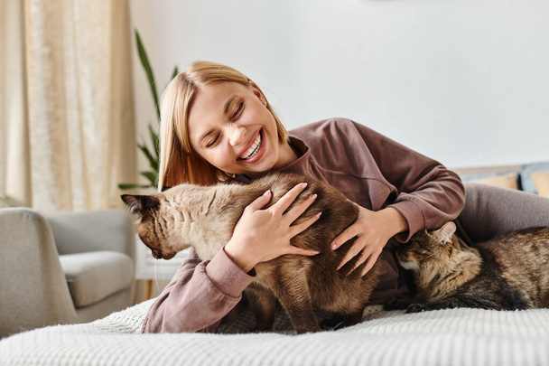 Atraktivní žena s krátkými vlasy pokojně dřímajícími na posteli, doprovázená dvěma hravými kočkami. - Fotografie, Obrázek
