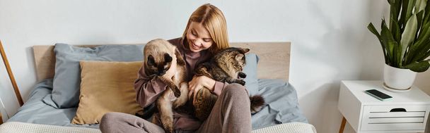 Una mujer con estilo se relaja en una cama con dos gatos, disfrutando de momentos de paz juntos en casa. - Foto, Imagen