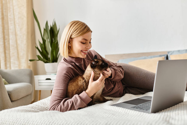 Mulher de cabelos curtos relaxando em uma cama com seu gato enquanto usa um laptop. - Foto, Imagem