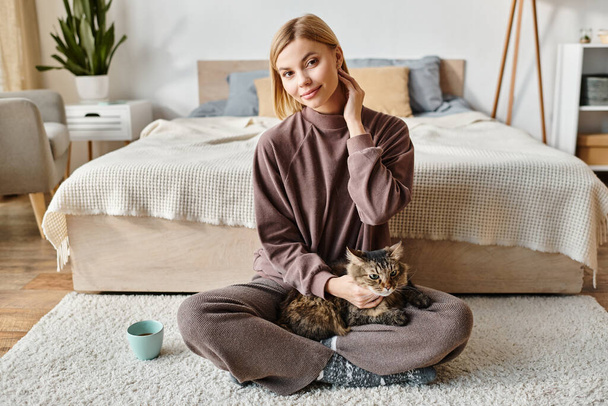 Egy rövid hajú nő békésen ül a padlón, a macskájával barátkozik az otthona kényelmében.. - Fotó, kép