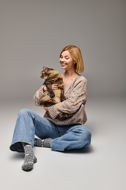 Kobieta z krótkimi włosami siedząca na podłodze, delikatnie trzymająca kota w spokojnej i intymnej chwili w domu. - Zdjęcie, obraz
