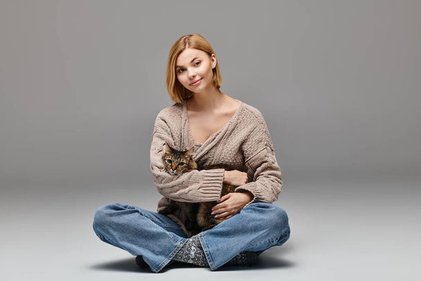 Una mujer atractiva con el pelo corto se sienta en el suelo, suavemente sosteniendo a un gato en su regazo, disfrutando de un momento tranquilo en casa. - Foto, imagen