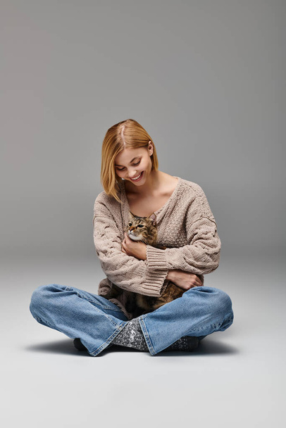 Uma mulher com cabelo curto senta-se no chão, segurando seu gato em um abraço amoroso em um ambiente acolhedor casa. - Foto, Imagem