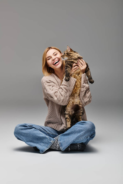 Una mujer con el pelo corto se sienta tranquilamente en el suelo, tiernamente sosteniendo a su preciado gato en sus brazos. - Foto, imagen