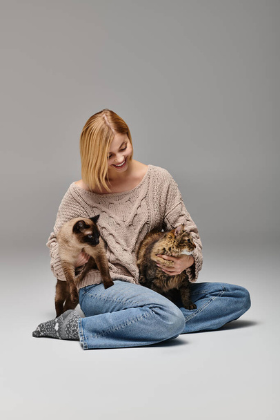 Žena s krátkými vlasy sedí na podlaze, něžně drží v náručí dvě kočky a předvádí klidný a klidný okamžik. - Fotografie, Obrázek