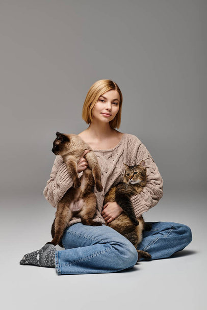 Egy rövid hajú nő ül a földön, két macskát ölel a karjaiban, egy békés és elégedett pillanatot testesítve meg otthon.. - Fotó, kép