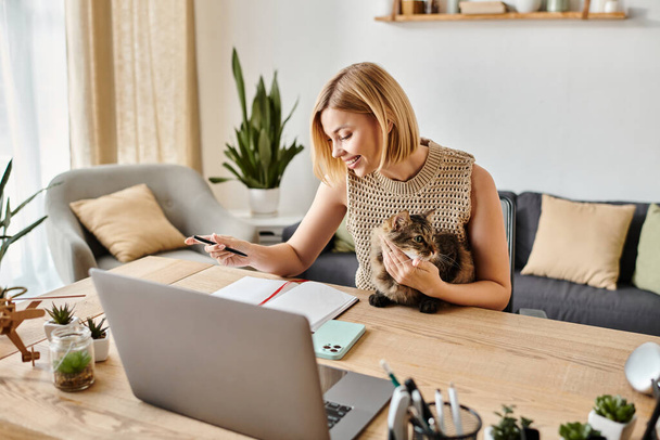 Una mujer elegante con el pelo corto trabaja en su computadora portátil en una mesa mientras su compañero felino peludo se sienta a su lado. - Foto, imagen