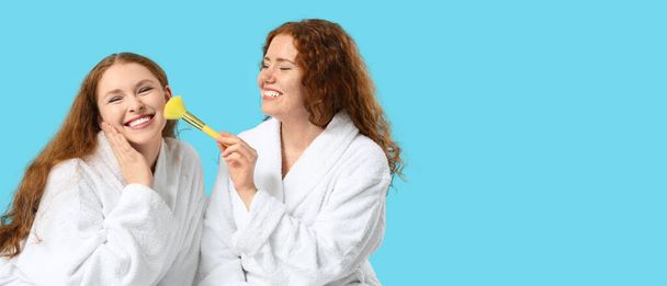 Bornozlu ve mavi arka planda makyaj fırçası olan mutlu kızıl kadınlar. - Fotoğraf, Görsel