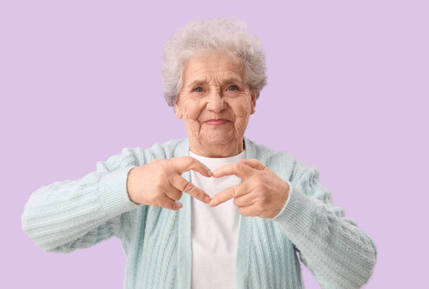 Старшая женщина делает сердце своими руками на сиреневом фоне - Фото, изображение