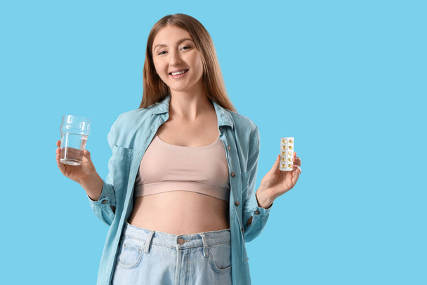Młoda kobieta w ciąży z tabletkami i szklanką wody na niebieskim tle - Zdjęcie, obraz