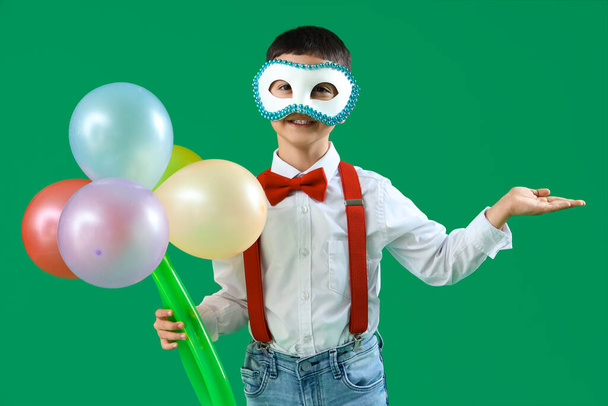 Grappig jongetje in kostuum en carnaval masker met boeket gemaakt van ballonnen tonen iets op groene achtergrond - Foto, afbeelding