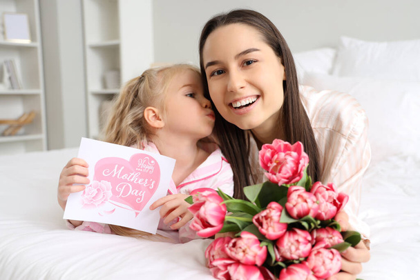 Niña felicitando a su madre feliz con ramo de hermosas flores y tarjeta de felicitación en el dormitorio. Celebración del Día de la Madre - Foto, imagen