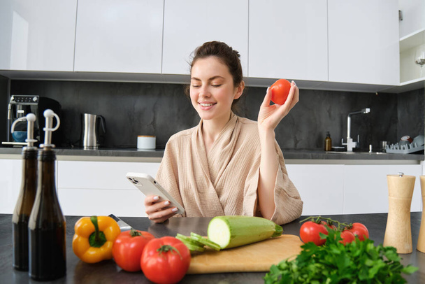 Imagen de una joven hermosa mujer, sosteniendo tomate, sentado en la cocina con teléfono inteligente, tabla de cortar y verduras en el mostrador, cocina de alimentos, compra de comestibles para su receta, utilizando la aplicación de teléfono móvil. - Foto, Imagen