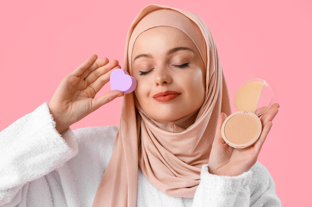 Hermosa mujer musulmana en hijab con esponja en forma de corazón y maquillaje en polvo sobre fondo rosa, primer plano - Foto, Imagen
