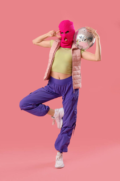 Beautiful young stylish woman in balaclava with disco ball showing victory gesture on pink background - Photo, Image