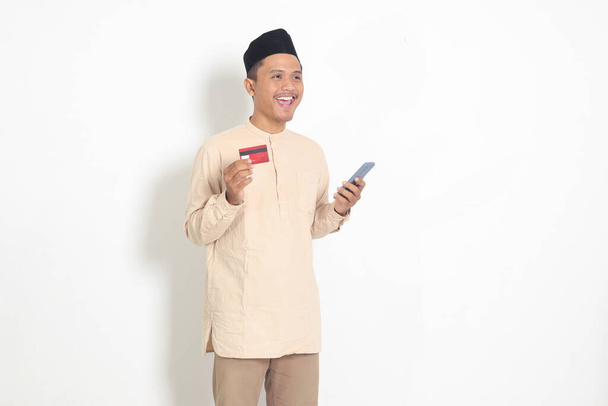 Elinde cep telefonu olan ve kredi kartı sunan Koko tişörtlü yakışıklı Asyalı Müslüman adamın portresi. Beyaz arkaplanda izole edilmiş resim - Fotoğraf, Görsel